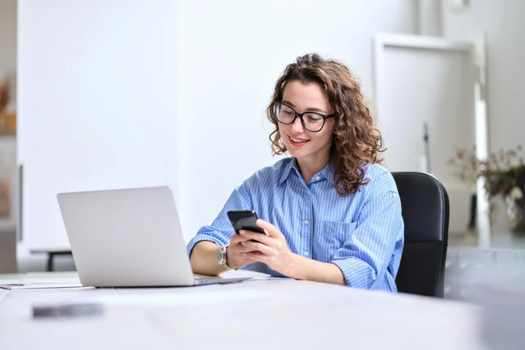 Happy woman sitting at desk with laptop using mobile phone to make an NDIS referral